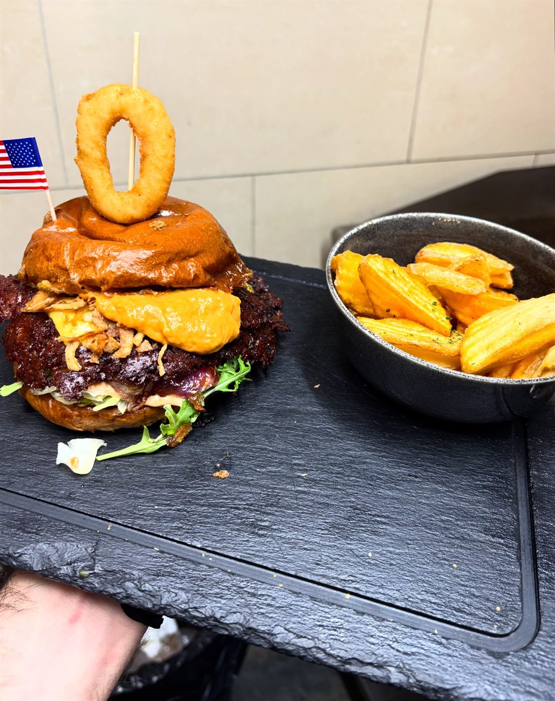 BBQ Burger mit Onion Ring obendrauf, serviert auf Schieferplatte mit Kartoffel-Wedges